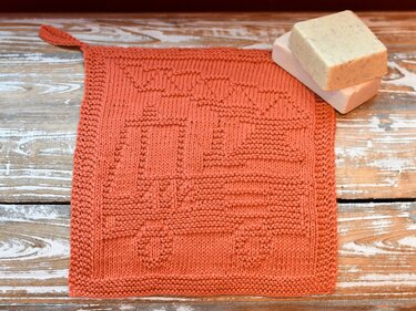 Quadratischer rostrot gestrickter Waschlappen mit erhabenem WASCH-Muster und Aufhängeschlaufe, neben zwei Seifenstücken auf Holz.