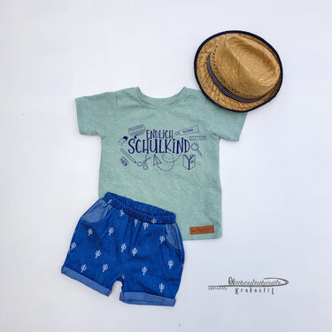 Hellgrünes Kinder-T-Shirt mit dunkelblauem Schriftzug 'Endlich Schulkind', daneben blaue Shorts mit Kaktusmuster und ein Strohhut auf wei
_em Hintergrund