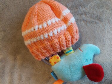 Orange Babymütze mit weißen Punkten neben türkisfarbenem Plüschtier auf weichem Untergrund