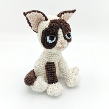 Sitting cream and brown amigurumi cat with pale blue stitched eyes on white background