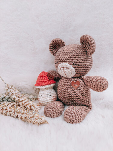 Brauner gehäkelter Amigurumi-Teddy mit Herz-Knopf neben kleinem Häkelpilz mit roter Kappe auf flauschigem Untergrund