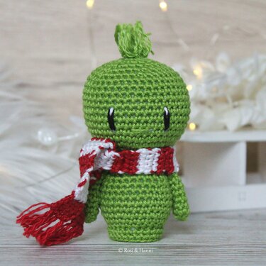 Grünes gehäkeltes Amigurumi mit rot-weiß gestreiftem Schal und Quaste auf dem Kopf vor hellem Hintergrund mit Lichterkette