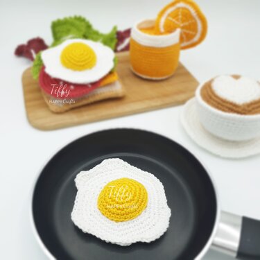 Gehäkeltes Spiegelei in einer schwarzen Pfanne, dahinter gehäkeltes Sandwich, Glas Orangensaft und Kaffeetasse auf hellem Hintergrund.