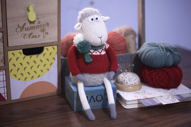 Knitted sheep amigurumi in red sweater and green scarf sitting on a box next to yarn and a pincushion