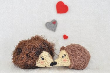 Zwei gehäkelte Igel stoßen mit den Nasen aneinander, vor roten und grauen Filzherzen auf weißem Hintergrund.