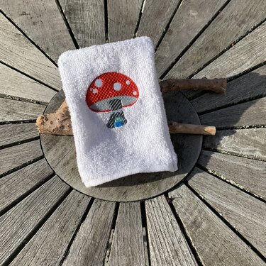 Nahaufnahme Stickmotiv Fliegenpilz mit roter Kappe, grauem Stiel und blauem Detail auf weißem Frottier
