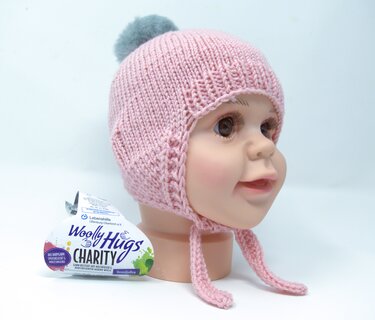 Rosa gestrickte Babymütze mit Ohrenklappen, Bindebändern und grauem Pompon auf einem Puppenkopf