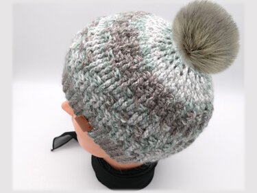 Gehäkelte Mütze in Grau, Weiß und Mint mit Bommel und kleinem braunen Label auf einem Mannequinkopf.