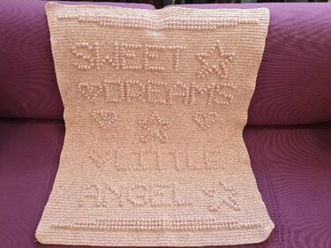 Zartrosa handgehäkelte Babydecke mit erhabenem Schriftzug „Sweet Dreams Little Angel“ und Stern‑ und Herzreliefs auf violettem Sofa.