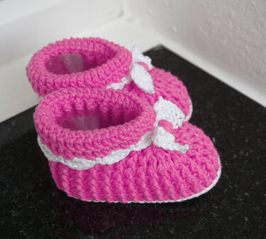 Pair of pink crocheted baby booties with white trim and bows on a dark surface