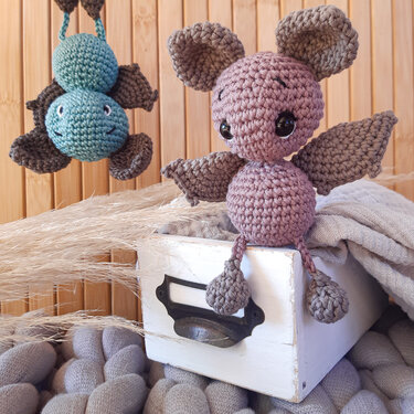 Zwei gehäkelte Amigurumi-Fledermäuse in Rosé und Türkis, eine sitzend auf einer weißen Schublade, eine hängend