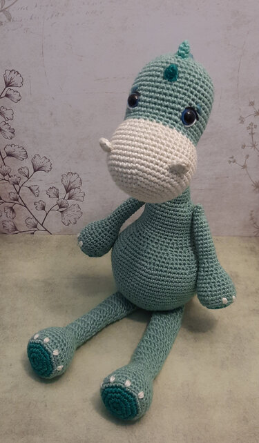 Gehäkelter Amigurumi‑Dino in Türkis und Weiß vor hellem Hintergrund mit floralem Muster