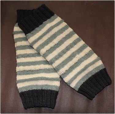 Zwei gestrickte Beinstulpen mit horizontalen Streifen in Dunkelgrau, Hellgrau und Creme auf braunem Untergrund.