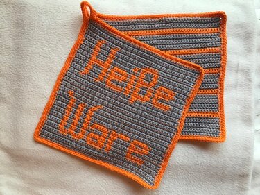 Zwei gehäkelte Topflappen in Grau und Orange; einer mit Schrift 'Heiße Ware', der andere gestreift