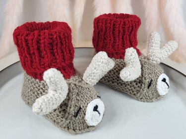 Gestrickte Rentier‑Babyschuhe mit roten Umschlagbündchen auf hellem Hintergrund
