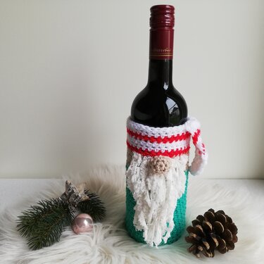 Weinflasche mit gehäkeltem Wichtel‑Überzug: grüner Körper, beige Mütze, weißer Zottelbart, kleine Nase und rot‑weiß gestreifter Schal. Dazu Tannenzweig, Christbaumkugel und Kiefernzapfen auf weißem flauschigem Untergrund.
