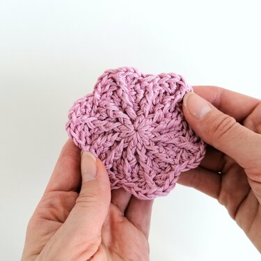 Hände halten ein rosa gehäkeltes Blütenmotiv mit sechs Blütenblättern