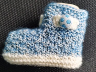 Gestrickter blau-weiß melierter Babyschuh mit weißer Kante und Fußballknopf auf dunklem Hintergrund