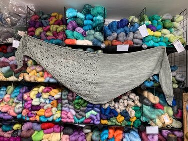 Hellgraues gestricktes Dreieckstuch mit Ajourmuster vor Regal mit bunten Garnen