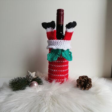Flasche mit gehäkelter Weihnachts-Hülle und Santa-Beinen, daneben Tannenzweig und Zapfen
