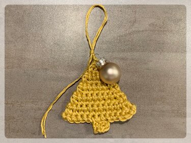 Nahaufnahme eines gelb gehäkelten Weihnachtsbaum-Anhängers mit einer matten goldfarbenen Kugel und einer Aufhängeschlaufe auf grauem Untergrund.