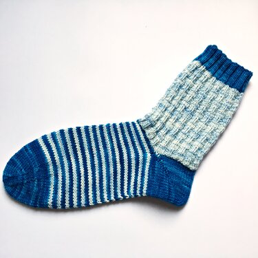 Gestrickte Socke in Blau und Weiß mit quer gestreiftem Fuß und geripptem Schaft auf weißem Hintergrund.