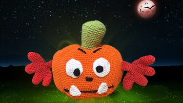 Gehäkelter orangefarbener Kürbis-Amigurumi mit großen Augen, spitzen Zähnen und roten Armen vor Nachthimmel mit rotem Mond und Fledermäusen.