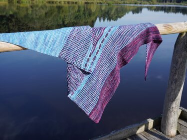 Gestricktes Dreieckstuch in Blau- und Violetttönen mit Loch- und Rippenmuster, über einem Holzgeländer am See drapiert.