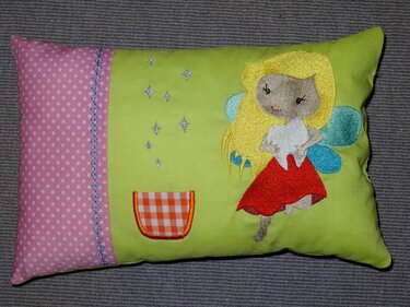 Stickdatei Zahnfee mit blonden Haaren, farbigen Flügeln, kariertem Täschchen und Sternen, auf einem Kinderkissen.