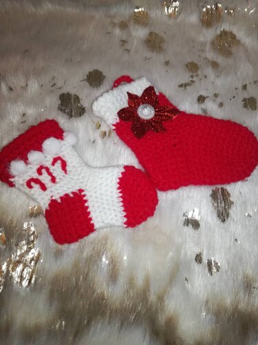 Zwei gehäkelte Weihnachtssocken rot-weiß, eine mit Pompons und roten Ornamenten, die andere mit glitzernder Blumenapplikation auf hellem, goldgetupftem Stoff.