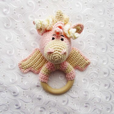 Pink and cream crocheted dragon-shaped baby rattle with a natural wooden ring on a white textured fabric.