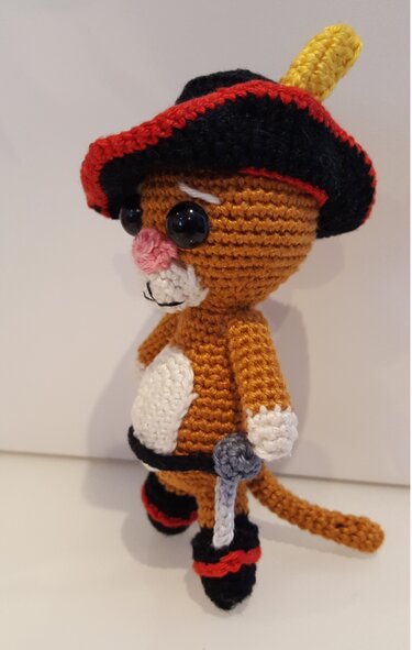 Gehäkelte braune Amigurumi-Katze mit schwarzem Hut, rotem Rand, gelber Feder und kleinem grauen Schwert