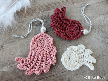 Drei gehäkelte Engelsflügel-Anhänger in Rosa, Bordeaux und Creme, zwei davon mit Aufhängekordeln.