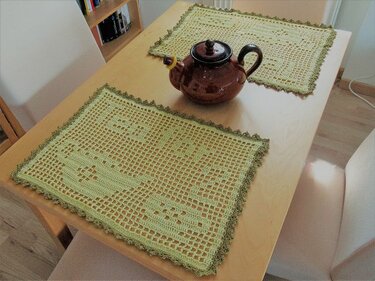 Zwei gehäkelte hellgrüne Platzsets mit dunklem Rand und eine braune Teekanne auf einem Holztisch.