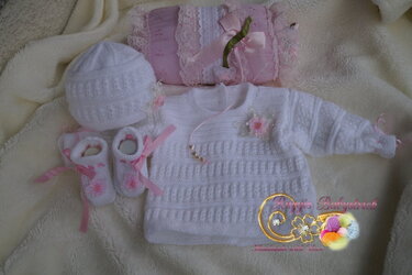 Weißes gestricktes Baby-Set mit Pullover, Mütze und Schühchen, rosa Schleifen und Blumenverzierung auf cremefarbener Decke