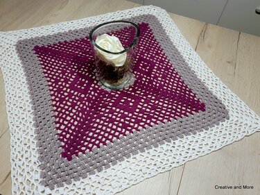 Quadratisches gehäkeltes Deckchen in dunklem Beerenton, Grau und Weiß auf hellem Holztisch mit Glasvase und weißer Rose
