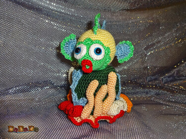 Buntes Amigurumi‑Meereswesen mit großen Augen, roten Lippen und tentakelähnlichen Häkelelementen auf glitzerndem Stoff.