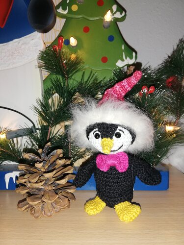Gehäkelter schwarzer Amigurumi-Pinguin mit rosa Weihnachtsmütze, weißem Fellrand, pinker Schleife sowie gelbem Schnabel und gelben Füßen neben Tannenzweigen und Zapfen