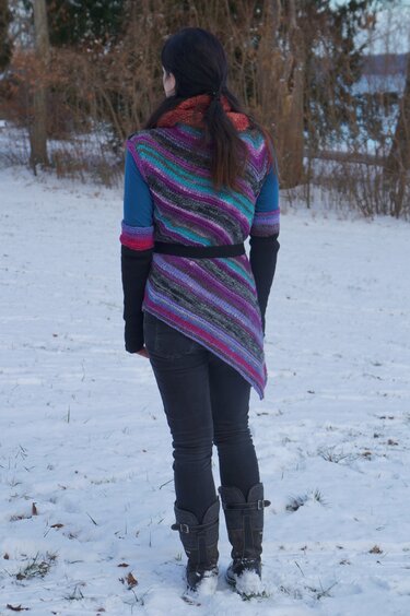 Rückansicht einer Person im Schnee mit mehrfarbigem gestrickten Wickelponcho, diagonalen Streifen, Gürtel und asymmetrischem Saum