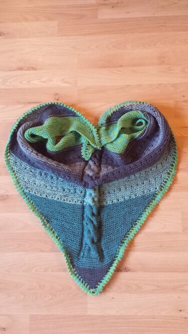 Gestricktes Dreieckstuch in Grün- und Blautönen mit zentralem Zopfmuster, Lochreihen und kontrastfarbiger Einfassung, herzförmig auf Holzboden drapiert.