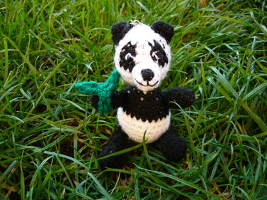 Gehäkelter schwarz-weißer Panda-Amigurumi sitzt im Gras mit kleinem grünen Blatt und Anhängerschlaufe