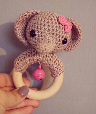 Gehäkelte Elefantenrassel mit rosa Schleife an hellem Holzring und rosa Glöckchen, von einer Hand gehalten.