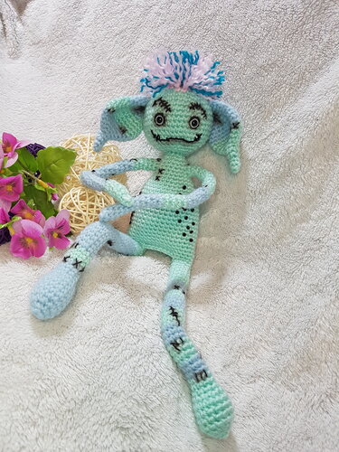 Gehäkeltes mintgrünes und hellblaues Amigurumi-Monster mit Pompom und großen Augen auf hellem Stoff