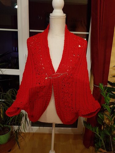 Roter gehäkelter Seelenwärmer mit Lochmuster auf Schneiderpuppe, vorne mit dekorativer Brosche befestigt.