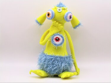 Gehäkeltes gelbes Amigurumi-Monster mit drei großen Augen und blauen flauschigen Akzenten, sitzend