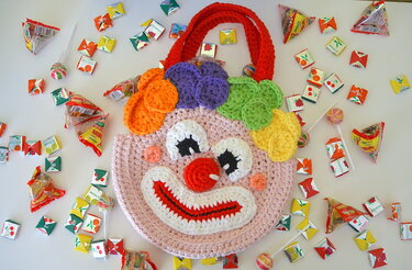 Handgehäkelte rosa Clown-Henkeltasche mit rotem Henkel und bunten Häkelapplikationen, umgeben von verpackten Süßigkeiten