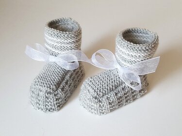Zwei handgestrickte hellgraue Babyschuhe mit feinen weißen Streifen und weißen Schleifen, Nahaufnahme des Strickmusters und der Verarbeitung.