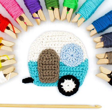 Häkelapplikation Wohnwagen türkis weiß mit schwarzem Rad, umgeben von buntem Garn und Häkelnadel