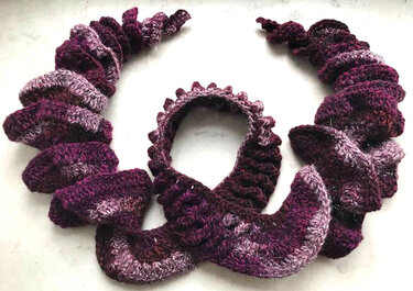 Gehäkelter Rüschenschal mit Spiralmuster in Violett‑ und Lilatönen, aufgelegt auf hellem Hintergrund