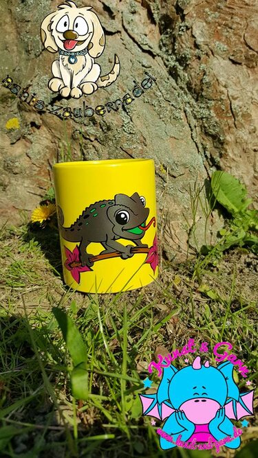 Gelbe Tasse mit stilisiertem Chamäleon auf Zweig und pinken Blüten, im Gras vor einem Baumstamm.